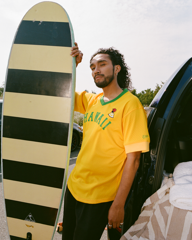 A man with long curly hair stands by an open car door holding a yellow and black surfboard, wearing an Ebbets Field Hawaii Islanders (PCL) Mesh V-Neck Jersey and looking at the camera with a relaxed expression.