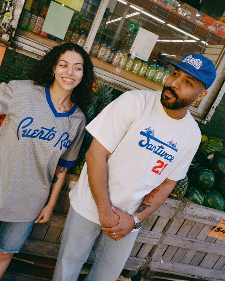 Two people stand outside a market. The woman wears a gray Puerto Rico jersey and denim shorts. The man, in light jeans, blue cap, and an Ebbets Field Santurce Cangrejeros Short Sleeve Tee nodding to baseball history, looks aside. Watermelons are stacked behind them.