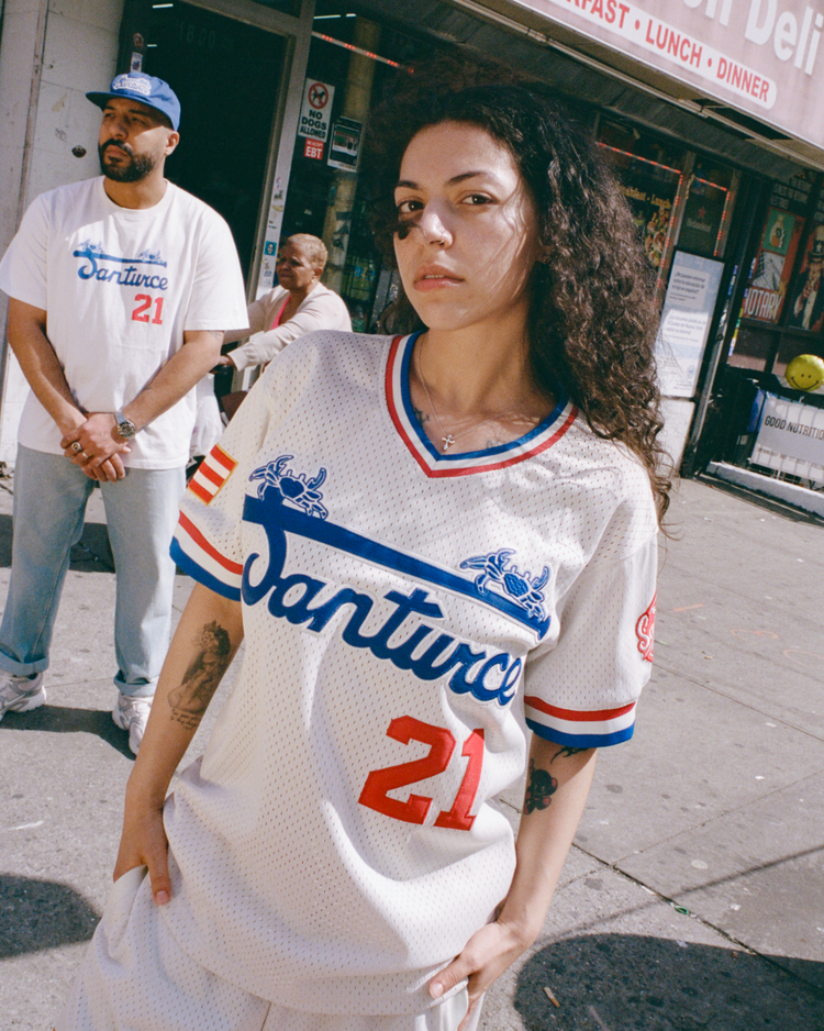 A woman wears an Ebbets Field Santurce Cangrejeros Mesh V-Neck Jersey as she stands confidently on a city sidewalk, with a man in a matching Puerto Rican baseball shirt and an older woman near a storefront behind her.