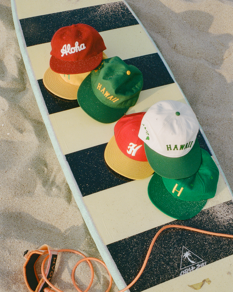 Five colorful wool baseball caps, including the EFF Hawaii Islanders 1961 Authentic Wool Ballcap, rest on a striped surfboard on the sand, with an orange leash attached.