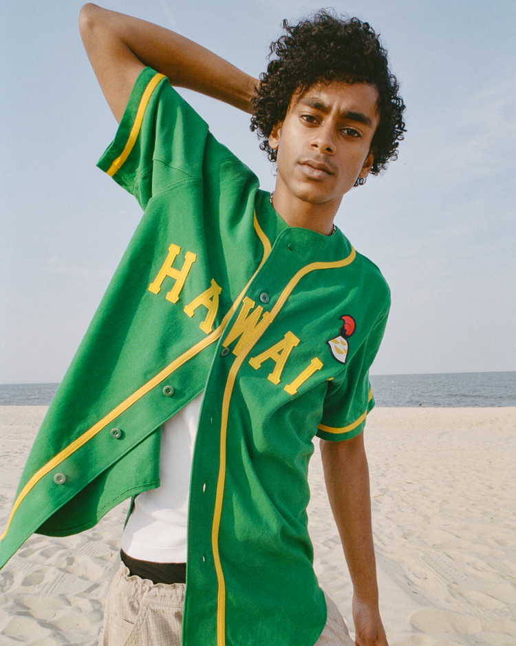 A person with curly hair stands on a sandy beach wearing the Ebbets Field Hawaii Islanders (PCL) Cotton Twill Baseball Jersey, layered over a white undershirt and paired with light pants, set against an ocean and sky Pacific Coast League backdrop.