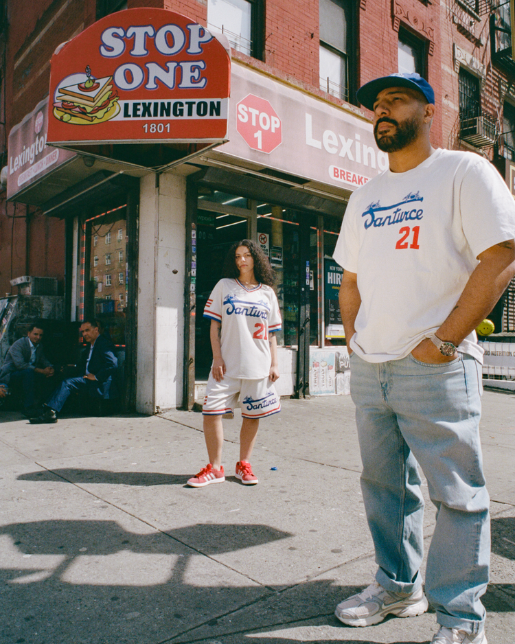 Two people in matching white Santurce Cangrejeros Short Sleeve Tees by Ebbets Field stand outside Stop One Lexington on a sunny day, while others sit near the entrance with brick buildings in the background.