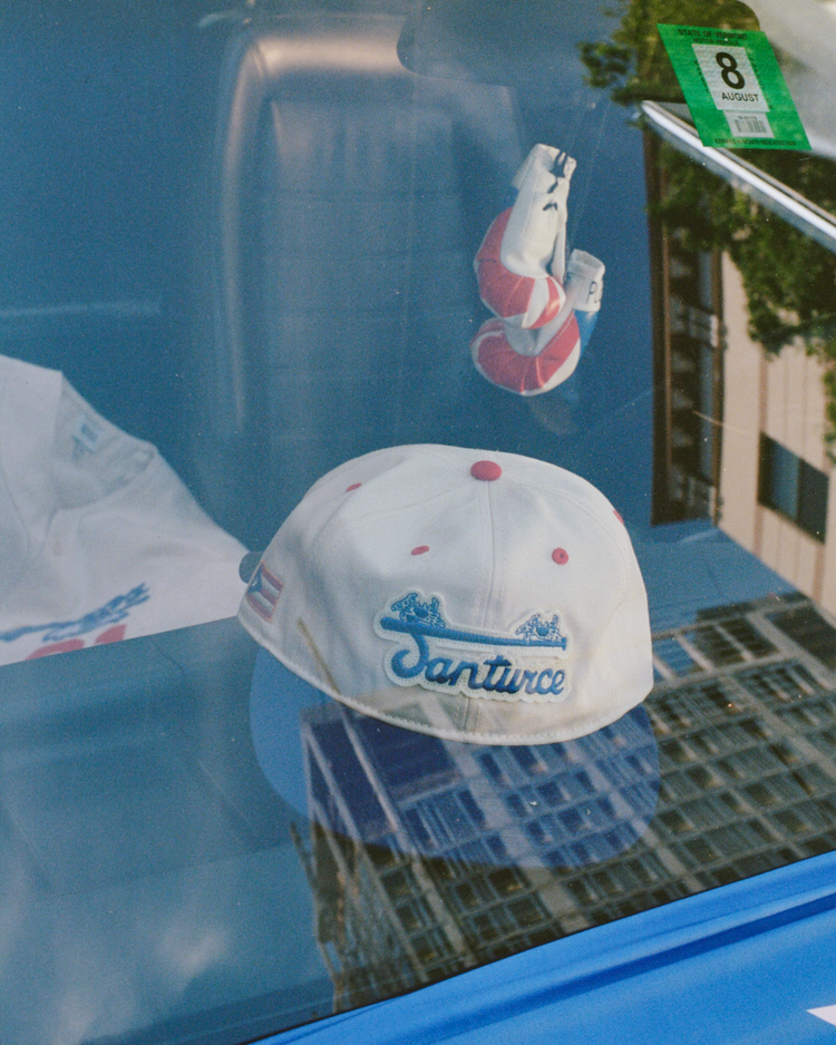 An Ebbets Field Santurce Cangrejeros Cotton Twill Ballcap with blue text and red dots rests on a car dashboard beside red-and-white boxing gloves and a green parking permit, city buildings reflecting Puerto Rican baseball spirit in the glass.