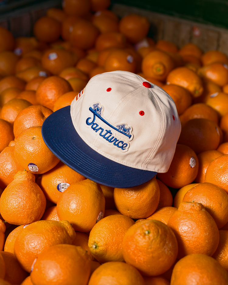 An Ebbets Field Santurce Cangrejeros Cotton Twill Ballcap in beige and navy sits atop a pile of oranges with stickers, brightly lit to celebrate Puerto Rican baseball and showcase both the cap and fresh fruit.