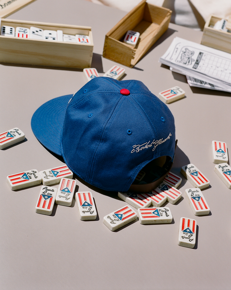 An Ebbets Field Santurce Cangrejeros Cotton Twill Ballcap sits with domino tiles showing the Puerto Rican flag, wooden boxes, and a score sheet—a nod to Puerto Rican baseball and Roberto Clemente.