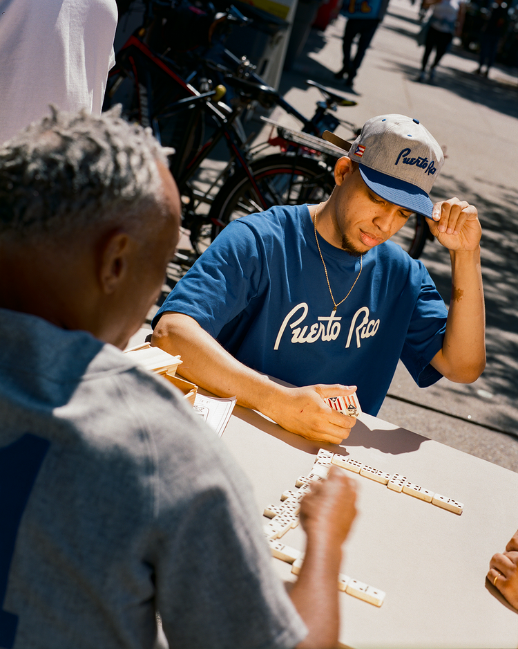 Two men play dominoes at an outdoor table. One wears the Ebbets Field Puerto Rico 1958 National Team Short Sleeve Tee and cap, focused on his tiles. Bicycles and people are visible in the sunny background.