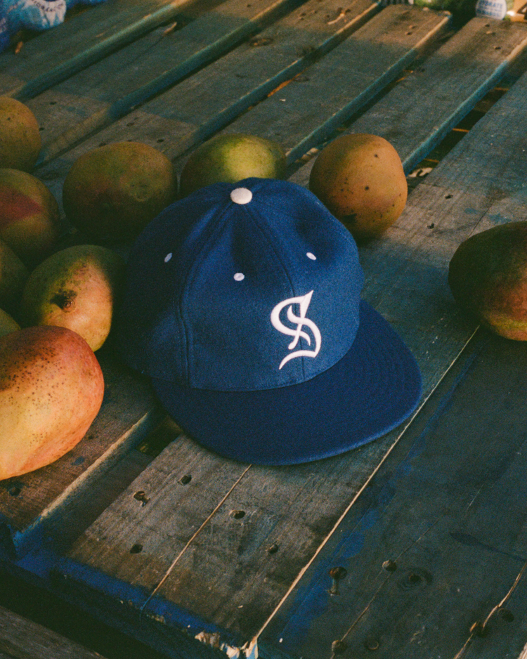 The EFF Santurce Cangrejeros 1954 Authentic Wool Ballcap, featuring a blue design and white embroidered logo, rests on a wooden surface surrounded by mangoes.