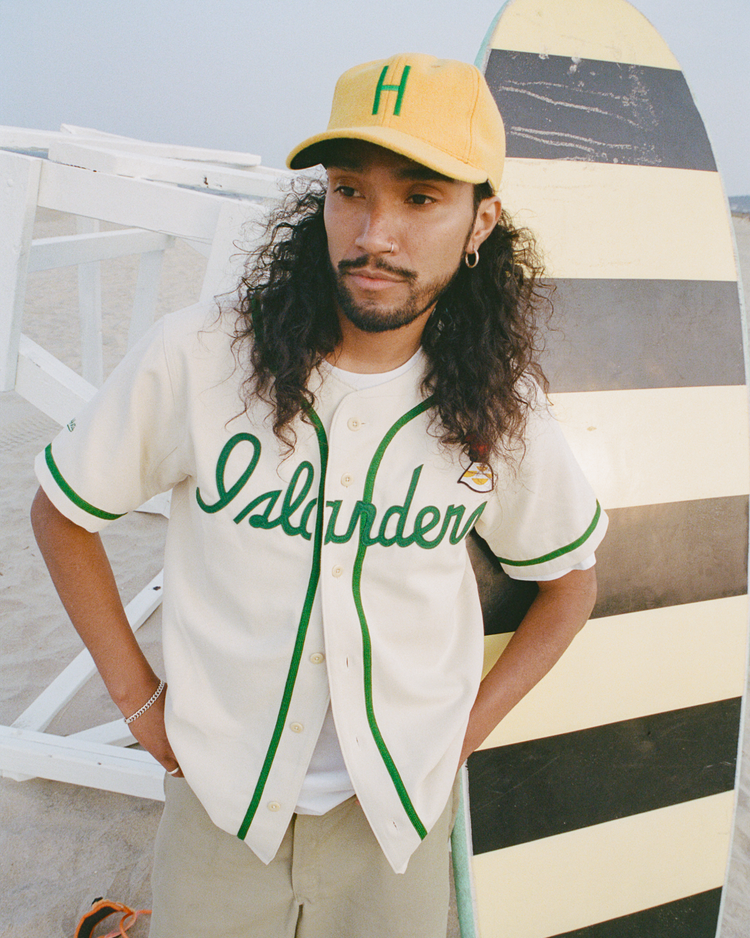 A man with long curly hair wears an EFF Hawaii Islanders 1961 Authentic Wool Ballcap and a white jersey with “Islanders” in green, standing on a sandy beach and leaning against a yellow-and-black striped surfboard.