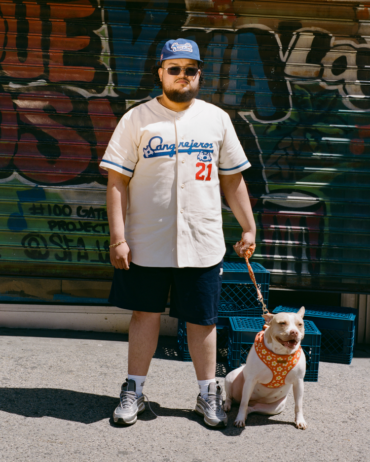 A man in sunglasses, shorts, a cap, and an Ebbets Field Santurce Cangrejeros 1954 Authentic Wool Home Baseball Jersey stands on the sidewalk with a leashed white dog. Behind them is a colorful graffiti-covered gate and stacked blue crates.