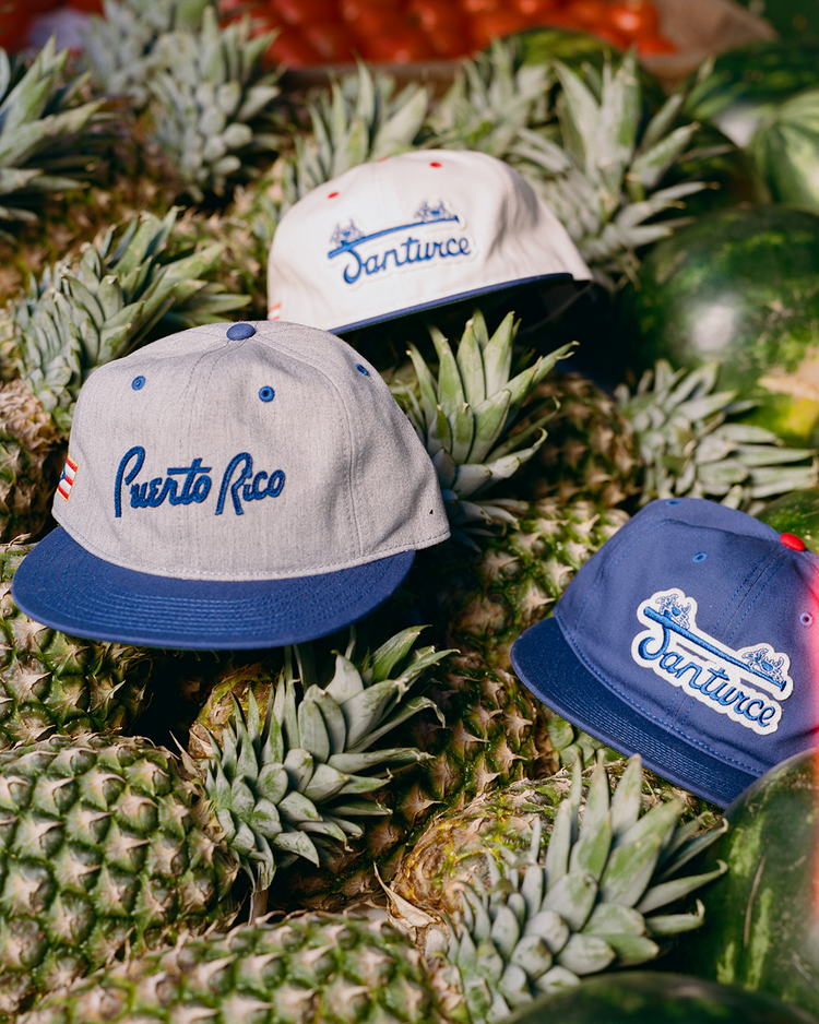 Three baseball caps, including an Ebbets Field Puerto Rico 1958 National Team Cotton Twill Ballcap, sit atop fresh pineapples. Two are blue and white, one is gray with a blue brim—celebrating Puerto Rican pride and baseball legends.