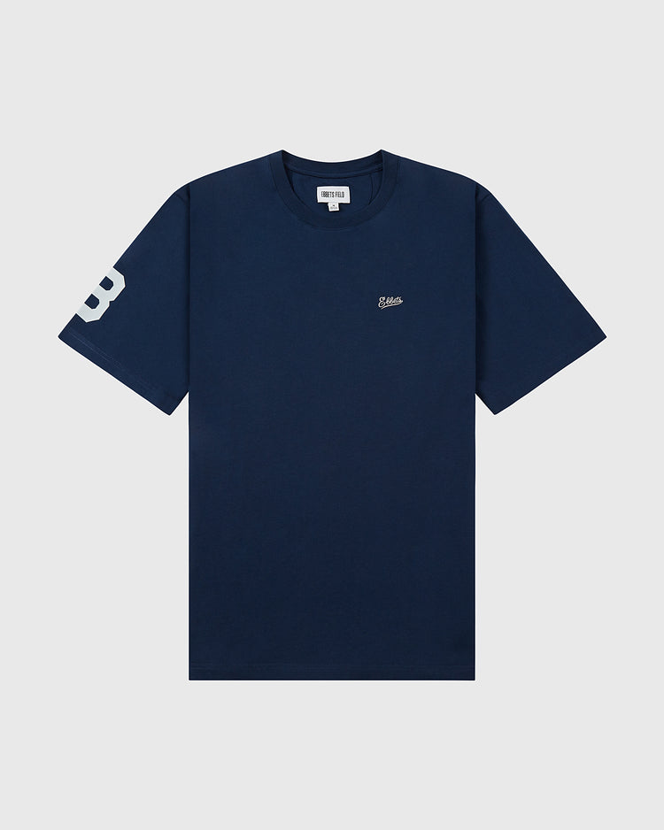 The Ebbets Field Brooklyn Bushwicks Ebbets Tee with Back Print is a navy blue short-sleeve t-shirt featuring a small white logo on the left chest and a large white B on the left sleeve, shown against a plain light background.