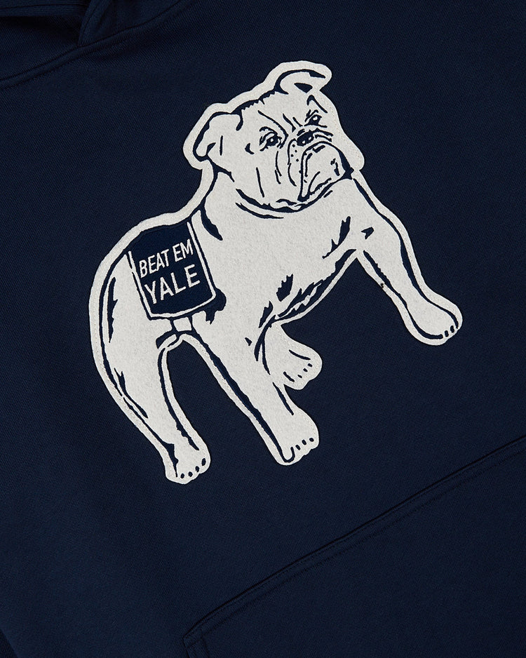 Illustration of a white bulldog in a BEAT EM YALE blanket appears on the navy YALE HOODIE by Ebbets Field, set against a dark blue fabric background.