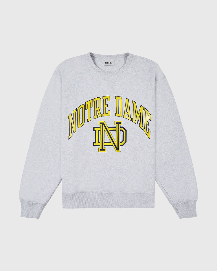 The NOTRE DAME CREWNECK SS H-GRY by Ebbets Field is a light gray crewneck sweatshirt featuring bold yellow NOTRE DAME lettering and a yellow ND monogram at the center.