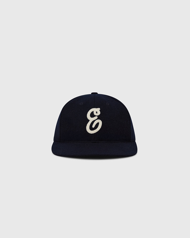 The Ebbets Felt 'E' Wool Ballcap by Ebbets Field is a navy wool broadcloth vintage cap with a white embroidered stylized 'E' on the front, shown against a light grey backdrop.