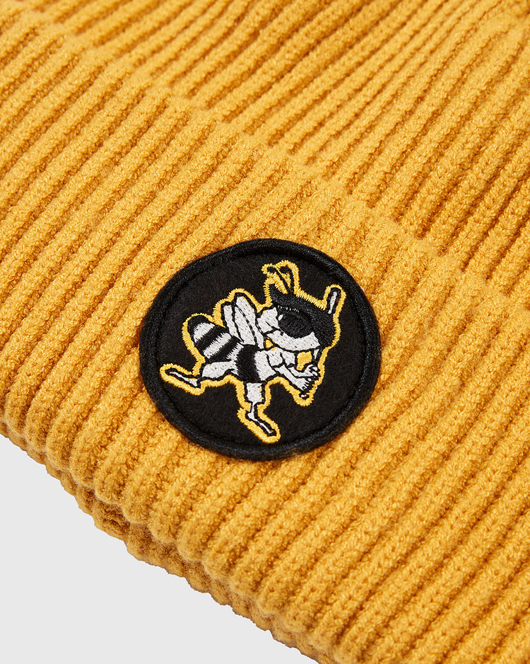 A close-up of the Ebbets Field Salt Lake Bees Patch Logo Ribbed Beanie in mustard yellow, featuring a circular black patch with an embroidered cartoon bee holding a bat and wearing sneakers, inspired by the Pacific Coast League team.