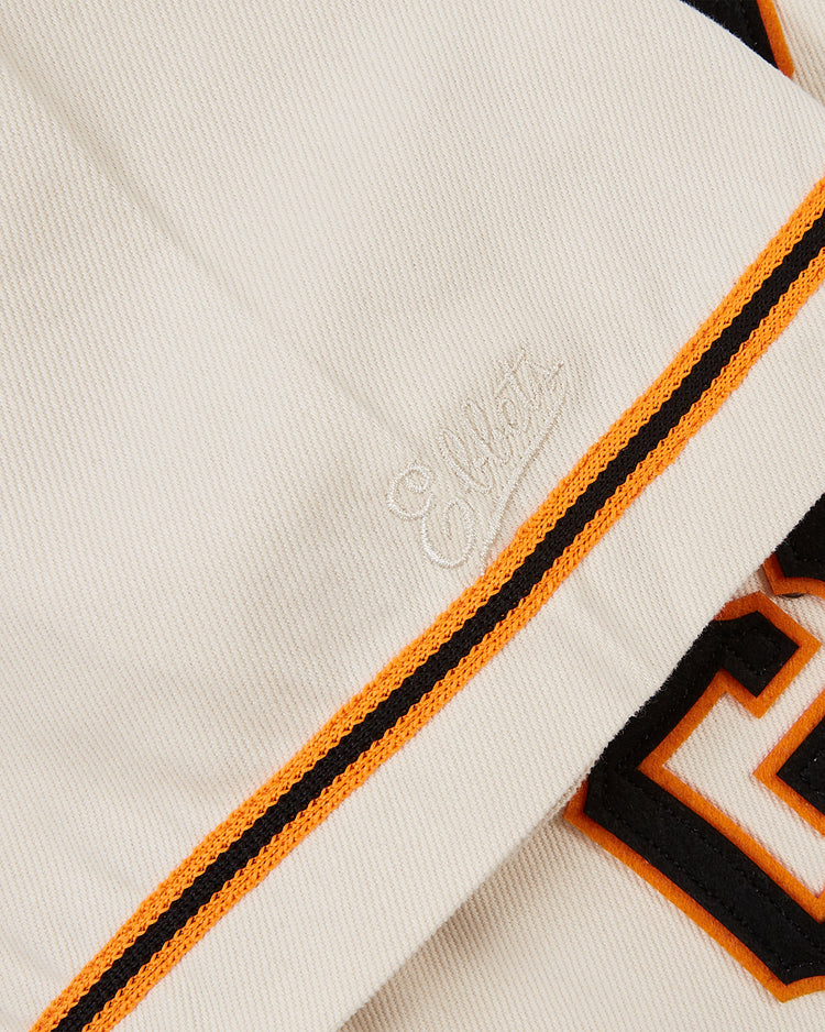 A close-up of the Tokyo Giants 1953 Cotton Twill Baseball Jersey by Ebbets Field, featuring cream-colored cotton twill with yellow and black stripes, a bold black and orange letter, and white script embroidery reminiscent of vintage baseball style.