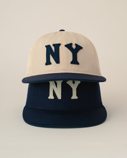 Two baseball caps stacked vertically; the top cap is cream-colored with large navy “NY” letters, and the bottom cap is navy with white “NY” letters. Both have flat brims and are displayed against a plain background.