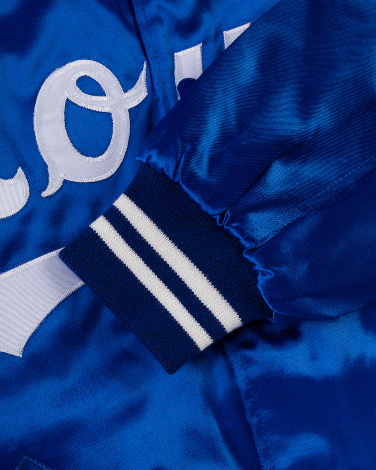 A close-up of an EFF Kansas City Royals 1969 Authentic All Satin Varsity Jacket sleeve, featuring shiny blue satin, a ribbed blue and white cuff, and part of a white embroidered letter on the fabric.