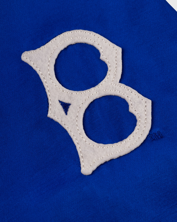 An EFF Brooklyn Dodgers 1945 Satin Windbreaker Jacket features a white “B” felt patch with two circular openings and visible edge stitching, sewn onto a bright blue fabric background.