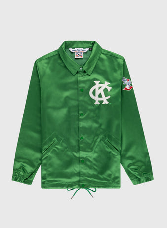 The EFF Kansas City Athletics 1963 Satin Windbreaker Jacket is a shiny green button-up with a white KC logo on the chest, colorful cartoon patch on the left sleeve, pointed collar, side pockets, and drawstring hem.