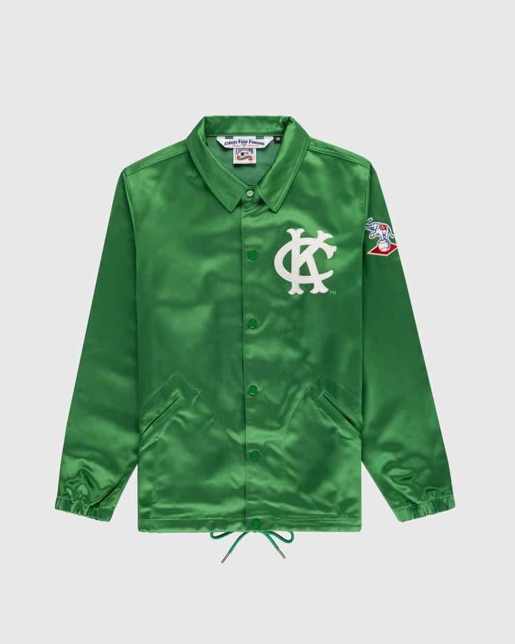 The EFF Kansas City Athletics 1963 Satin Windbreaker Jacket is a shiny green button-up with a white KC logo on the chest, colorful cartoon patch on the left sleeve, pointed collar, side pockets, and drawstring hem.