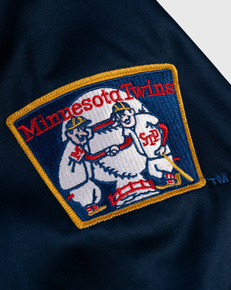 The EFF Minnesota Twins 1966 Satin Windbreaker Jacket features an embroidered patch of two baseball players shaking hands over a baseball, with red and yellow text on dark blue—an iconic vintage MLB look.
