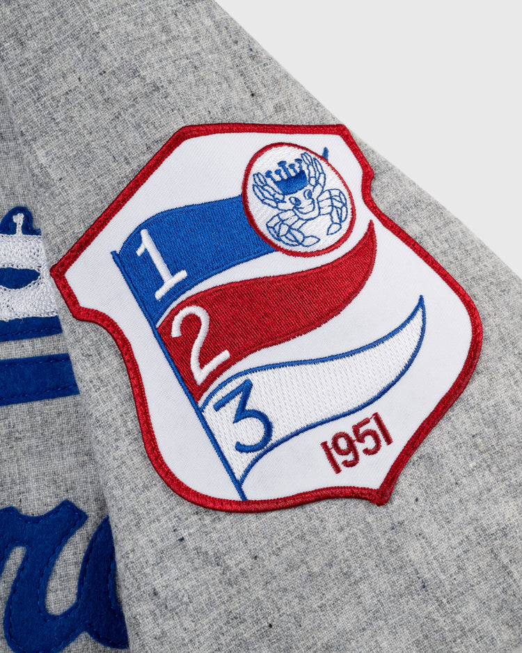 A close-up of gray wool fabric on the Ebbets Field Santurce Cangrejeros 1954 Authentic Road Jersey highlights a colorful embroidered patch with a red, blue, and white shield, numbers 1, 2, 3, a baseball glove and ball, and 1954.