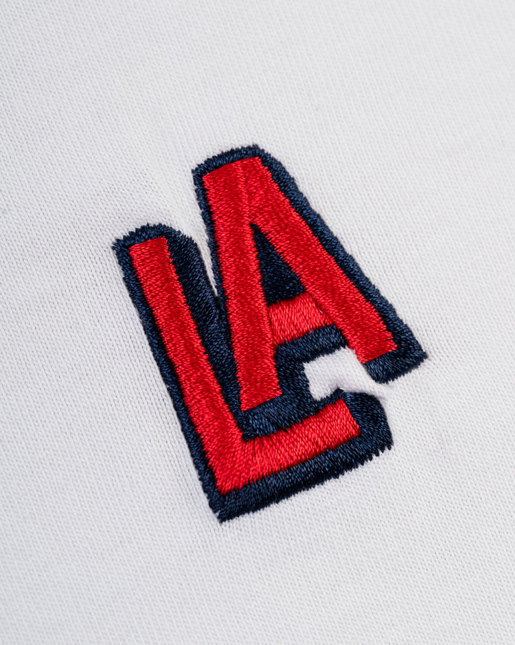 The Ebbets Field Los Angeles Angels (PCL) Embroidered ‘LA’ Short Sleeve Tee features a bold red and navy blue 1942 LA logo on white fabric, with overlapping letters, inspired by classic Ebbets tee design.