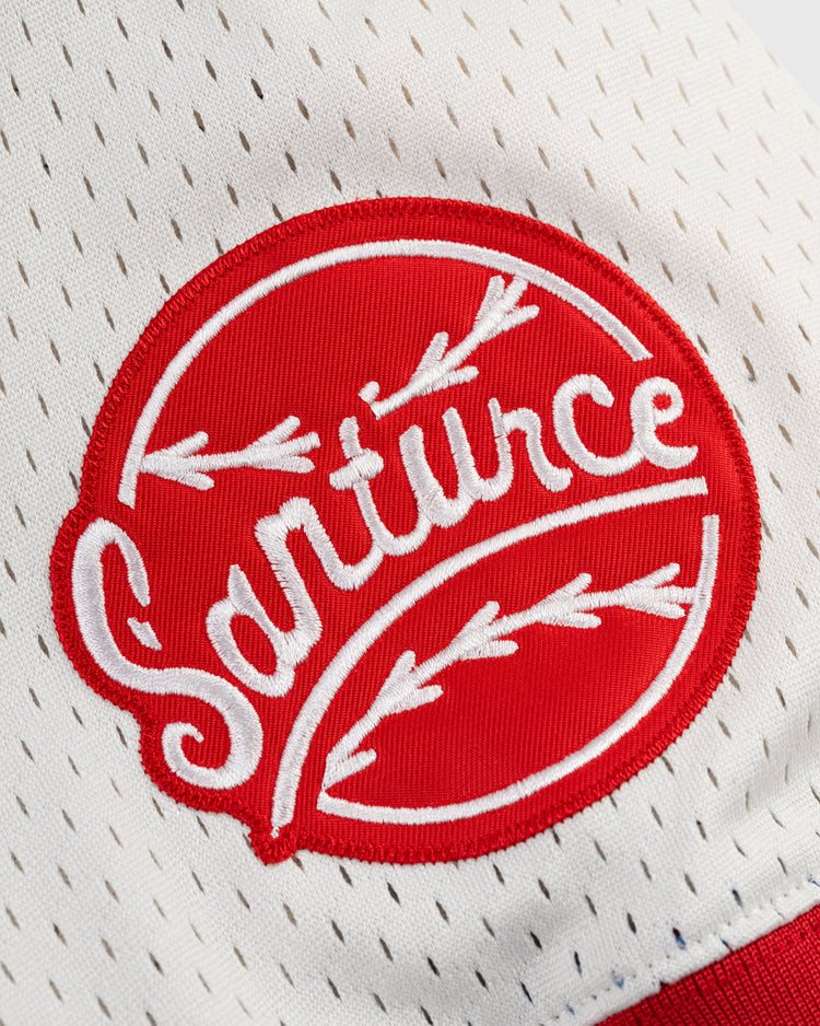 Close-up of the Ebbets Field Santurce Cangrejeros Mesh V-Neck Jersey, showing a red and white embroidered patch with Santurce in stylized letters and arrow designs on perforated white mesh fabric.