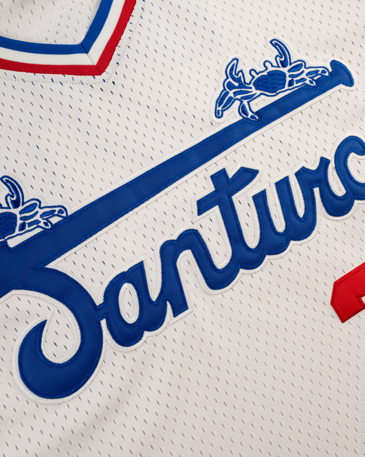 Close-up of the Ebbets Field Santurce Cangrejeros Mesh V-Neck Jersey in white with blue and red trim. “Santos” appears in blue across the front, with blue horse rider graphics above—honoring Puerto Rican baseball heritage.