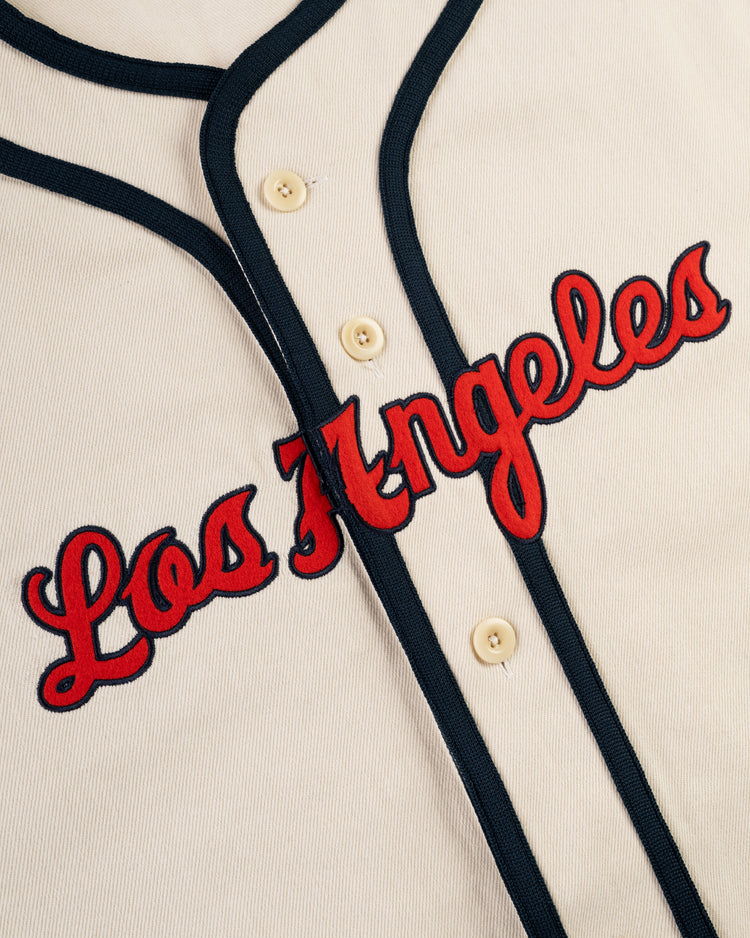A close-up of the Ebbets Field Los Angeles Angels (PCL) Cotton Twill Jersey shows dark blue trim, red script Los Angeles lettering across the chest, and four cream buttons, evoking classic Pacific Coast League style.