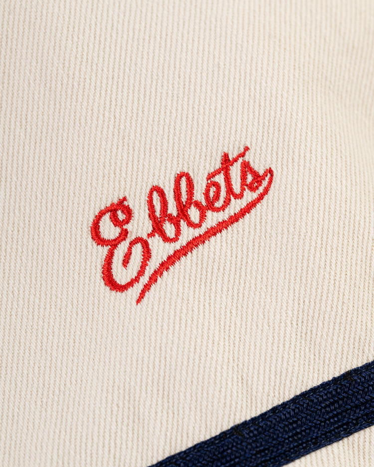 Close-up of cream-colored cotton twill from the Los Angeles Angels (PCL) jersey by Ebbets Field, featuring Ebbets in red cursive embroidery and a navy blue stripe, inspired by classic Pacific Coast League style.