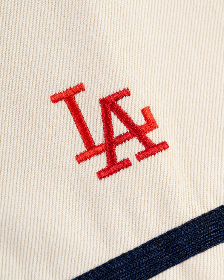 A close-up of the Ebbets Field Los Angeles Angels (PCL) Cotton Twill Jersey shows cream fabric with a red embroidered LA logo and a navy blue stripe, evoking classic Pacific Coast League baseball style.