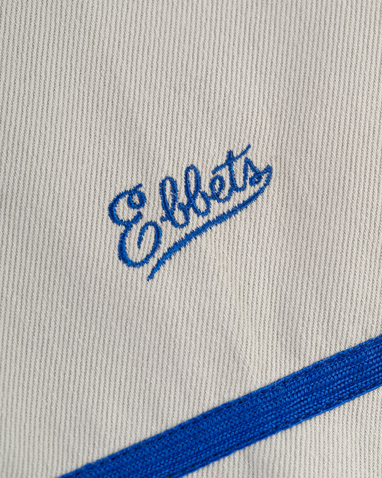 Close-up of white cotton twill fabric on the Ebbets Field Puerto Rico 1958 National Team Baseball Jersey, featuring blue cursive Ebbets embroidery and a diagonal blue stripe at the bottom right corner.