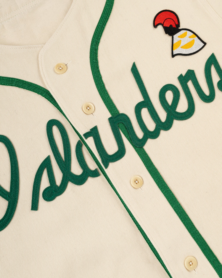 Close-up of the Ebbets Field Hawaii Islanders (PCL) Cotton Twill Baseball Jersey, featuring cream color, green trim, Islanders in green script, and a red-and-yellow bell patch near the shoulder—echoing the classic Hawaii Islanders style.