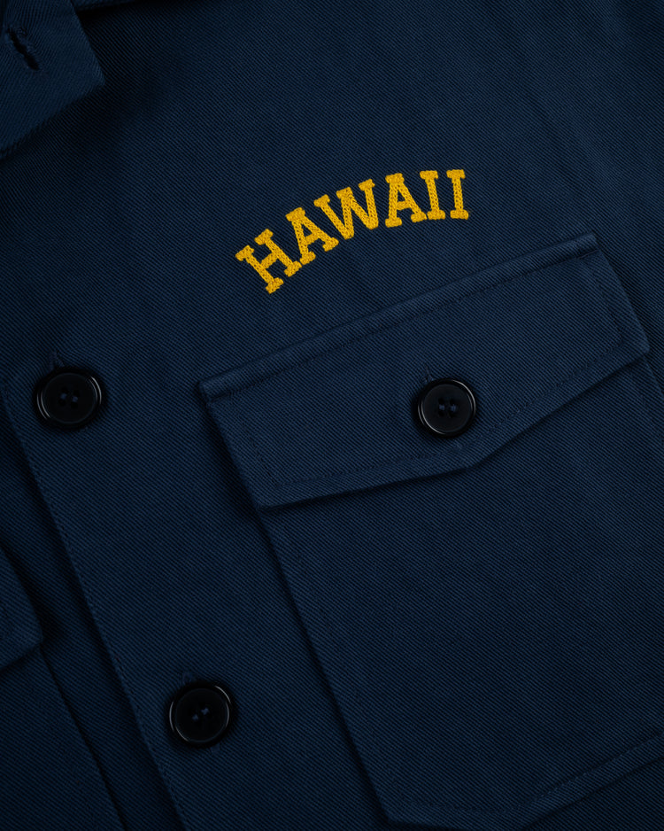 A close-up of the Ebbets Field Hawaii Islanders (PCL) Cotton Linen Grounds Crew Short Sleeve Workshirt in navy blue, featuring black buttons and HAWAII embroidered in yellow above the chest pocket.
