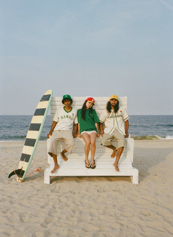 Three people wearing Hawaii-themed baseball shirts and caps sit by a white wooden wall at the beach, with a green surfboard and the ocean in the background. They look relaxed and casual.
