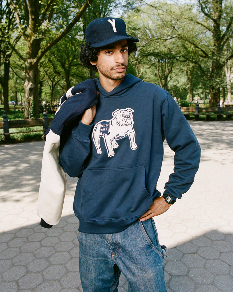 A man stands outdoors in a navy hoodie with a Yale Bulldogs Baseball 1948 Championship graphic, holding a jacket. He wears EFF’s Yale University 1948 Authentic Wool Ballcap with a Y, jeans, and a watch; trees and a park path are behind him.