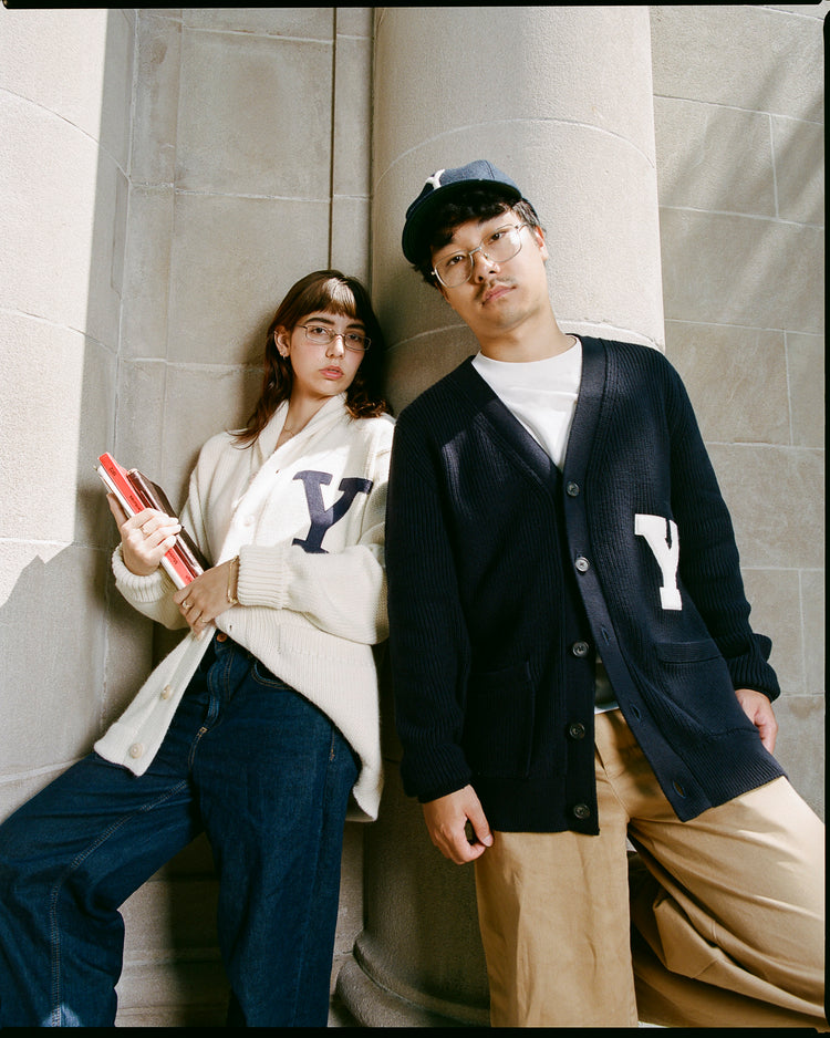 Two people lean against a stone column, both wearing EFF Yale University 1923 Wool Shawl Collar Sweaters with bold Y letters—one holding books, the other in a baseball cap and glasses, capturing a classic Yale vibe.