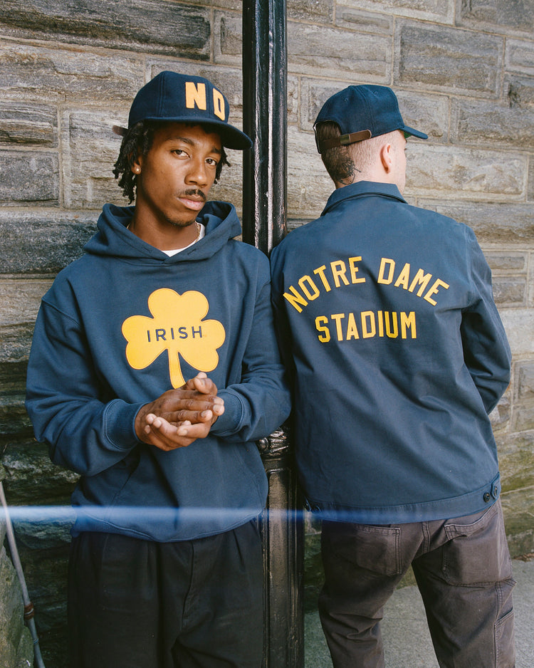Two people in navy outfits stand by a stone wall. One faces the camera wearing an Ebbets Field University of Notre Dame Heavyweight Cotton Terry Pullover Hoodie with a yellow shamrock and IRISH on the front.