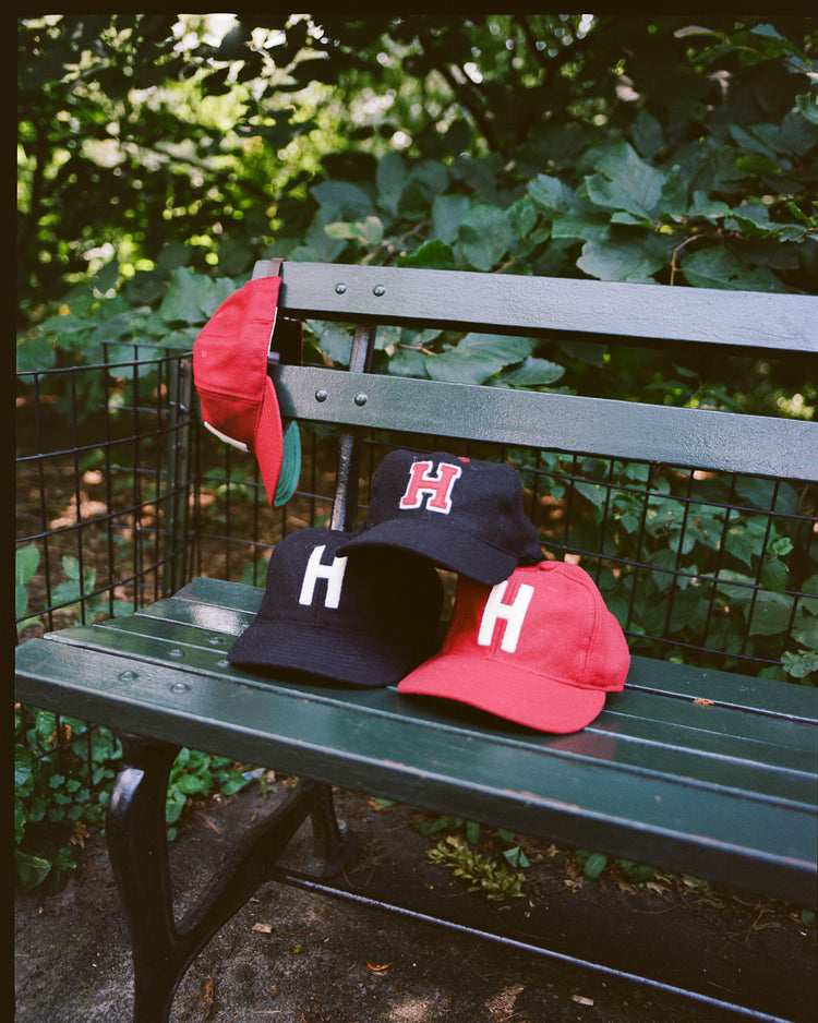 Four EFF Harvard University 1950 Authentic Wool Ballcaps—two black, two red—are displayed on a green park bench. One red cap hangs from the backrest, set among lush greenery.