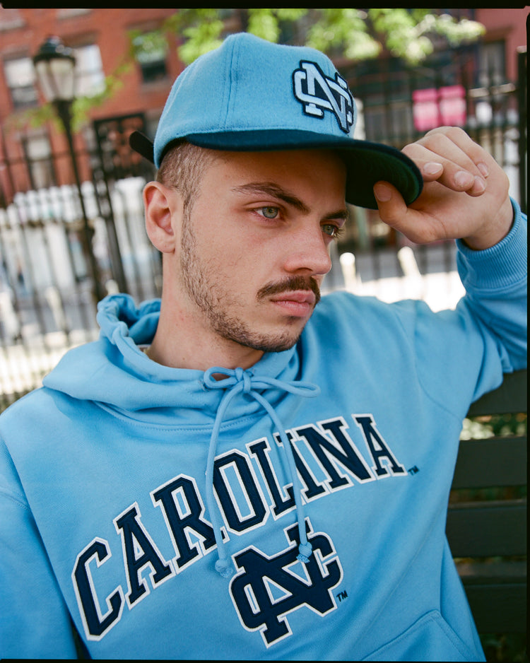 A young man wearing a blue Carolina hoodie and an EFF University of North Carolina 1983 Vintage Ballcap sits thoughtfully on a bench outdoors, with a blurred fence, trees, and a brick building in the background.