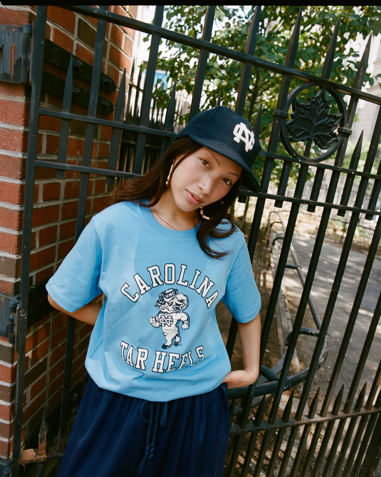 A woman in an Ebbets Field UNIVERSITY OF NORTH CAROLINA SS TEE BLUE and a navy cap leans against a black iron gate outdoors, with trees and a brick wall behind her.