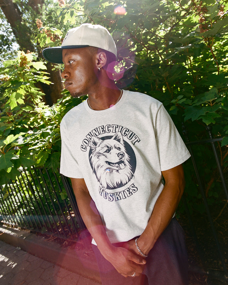 A man wearing the Ebbets Field UCONN SS TEE H-GRY stands outdoors by black railings and leafy green trees, looking to his left with sunlight streaming through the background.