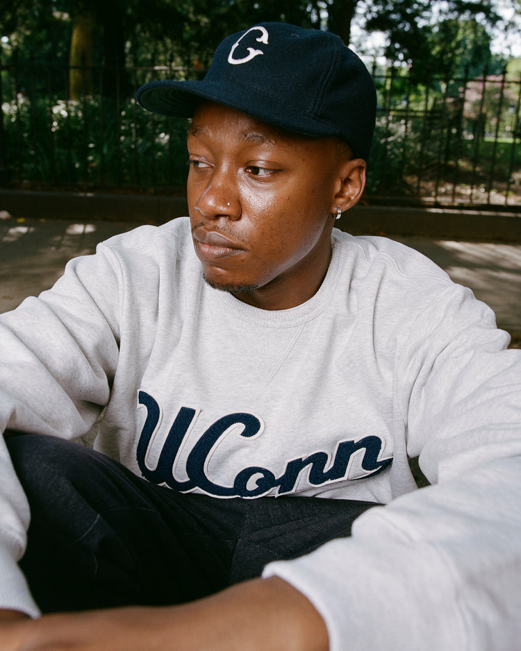 A young man in an EFF University of Connecticut 1957 Vintage Ballcap and a light gray UConn sweatshirt sits outdoors, gazing thoughtfully to the side, with trees and a metal fence blurred in the background.