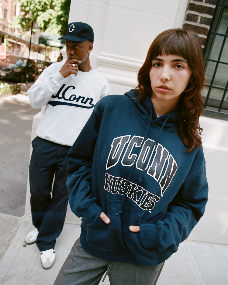 Two people stand on a sidewalk. The person in front wears an Ebbets Field UCONN HOODIE NAVY, while the person behind wears a white UConn sweatshirt, navy pants, and a baseball cap. Trees and buildings are visible in the background.