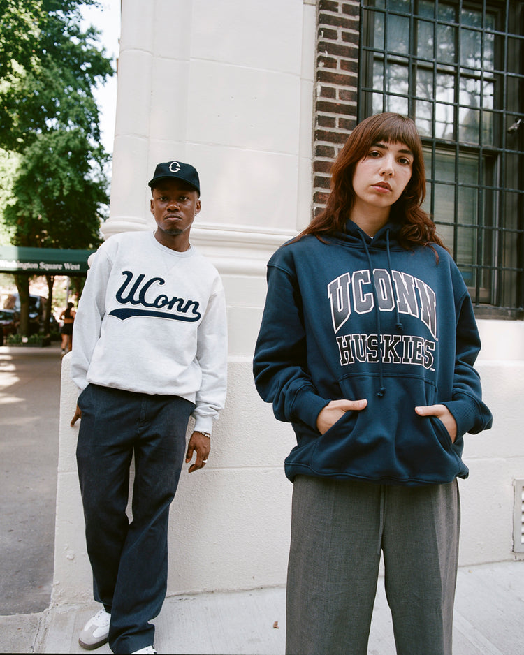 Two people stand on a city sidewalk; one wears an Ebbets Field UCONN CREWNECK SS H-GRY sweatshirt, the other sports a navy UConn Huskies hoodie and gray pants. A brick and stone building with greenery is in the background.