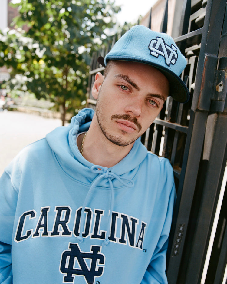 A man in an Ebbets Field UNIVERSITY OF NORTH CAROLINA HOODIE BLUE and matching cap stands by a metal fence, facing the camera with sunlit leaves in the background.