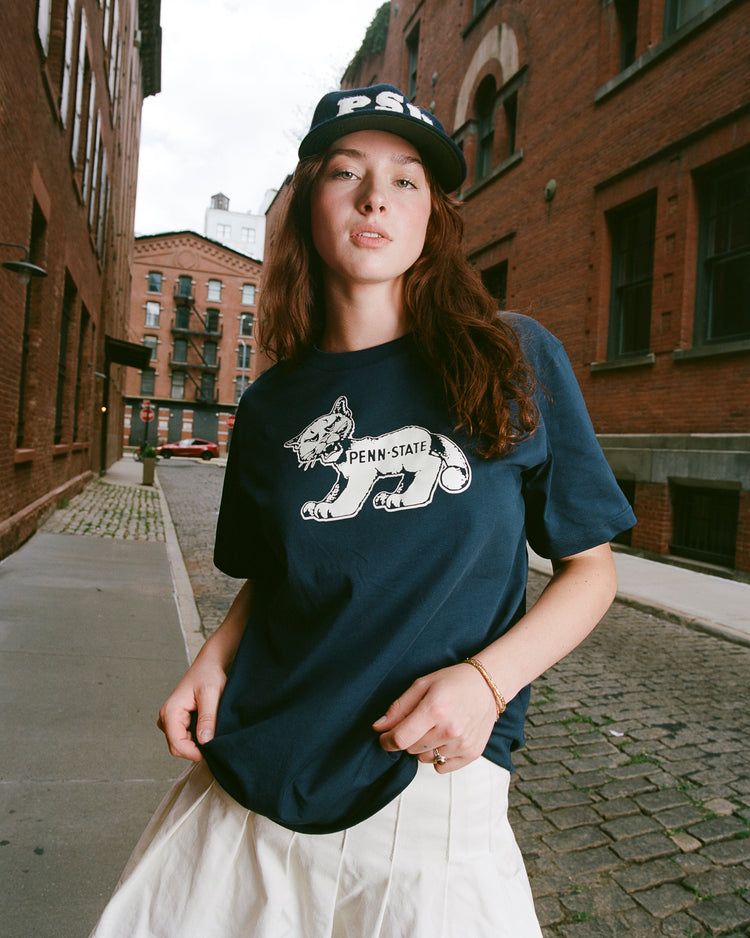 A woman stands on a cobblestone street between brick buildings, wearing an Ebbets Field PENN STATE SS TEE in navy, a matching PS cap, and a white skirt, looking confidently at the camera.
