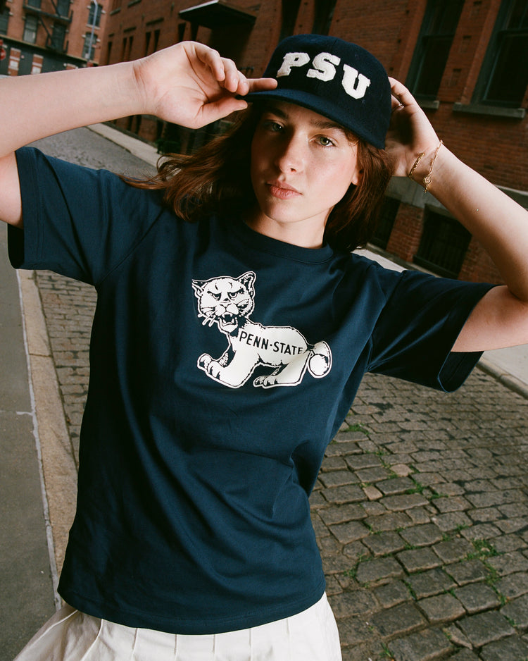 A person stands on a cobblestone street wearing a navy Penn State t-shirt with a cartoon lion and an Ebbets Field Penn State University Authentic Wool Ballcap, holding the brim with both hands. Brick buildings line the background.